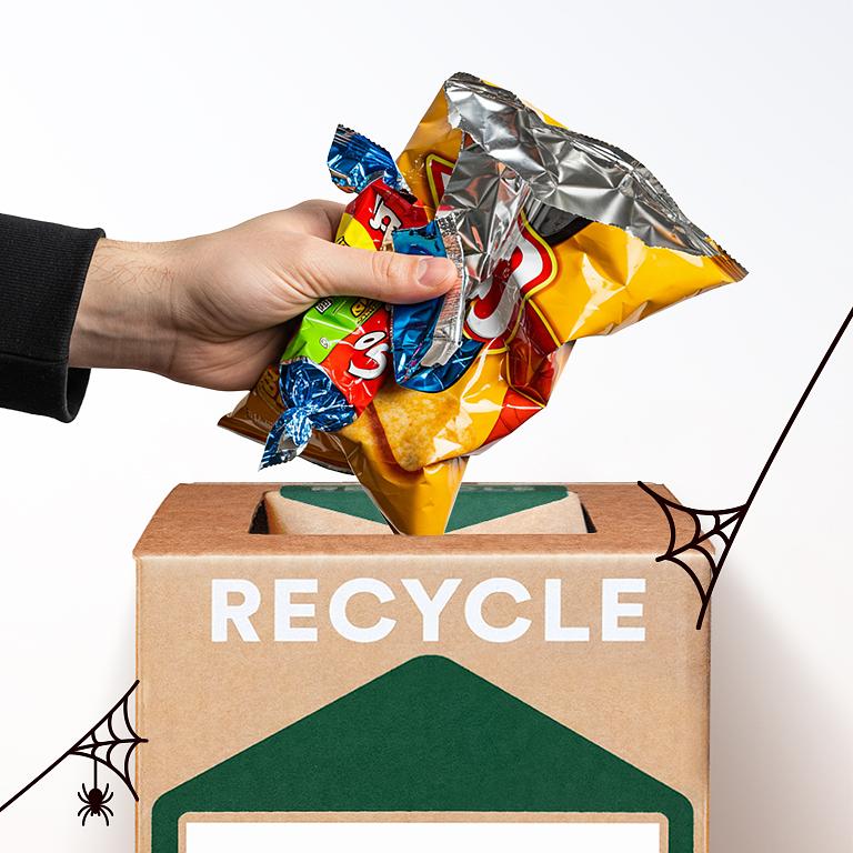 A hand places empty candy wrappers into a recycling bin decorated with drawn cobwebs.