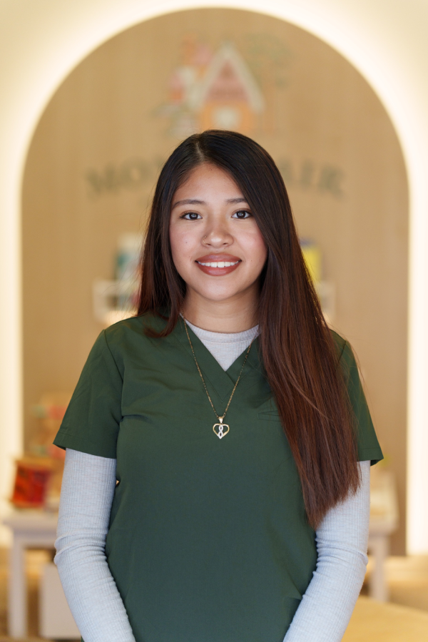 Smiling staff member with long straight hair and a heart-shaped necklace, wearing a green scrub top layered over a white long-sleeve, standing in front of a wooden archway with a "Montclair" sign.