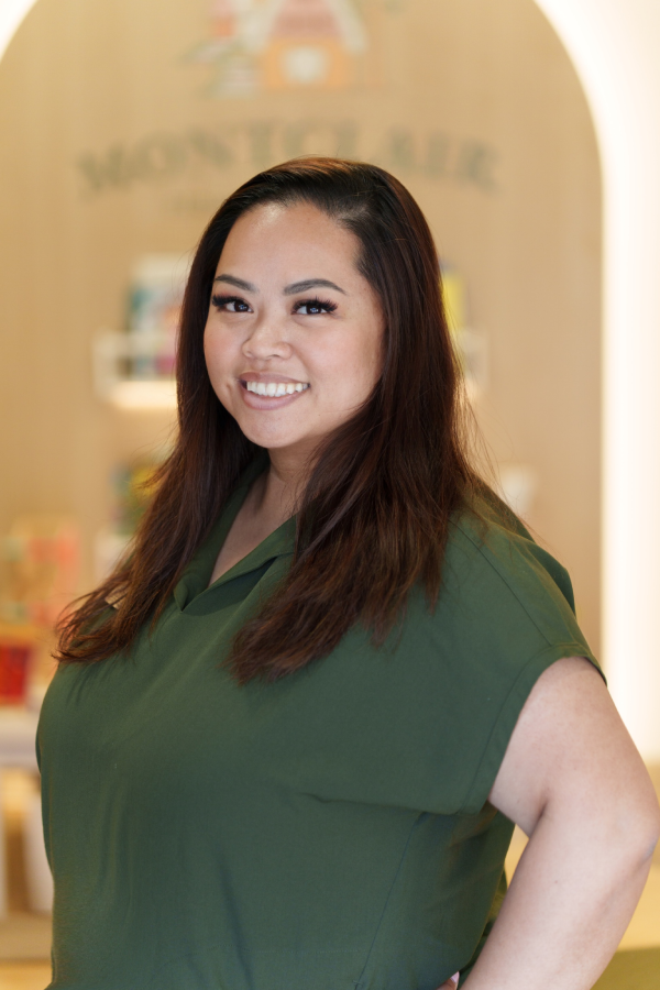 Smiling staff member with long dark hair, wearing a green top, standing in front of a wooden archway with a "Montclair" pediatric dental sign.
