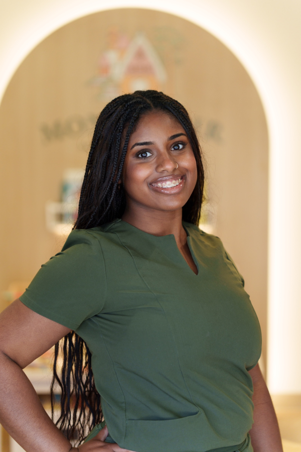 Smiling staff member with long braided hair wearing a green scrub top, standing in front of a wooden archway with a "Montclair" pediatric dental sign.