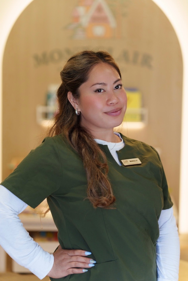 Staff member with a half-up hairstyle, wearing a green scrub top with a name badge reading "Marcela" over a white long-sleeve, standing with hand on hip in front of a wooden archway with a "Montclair" sign.