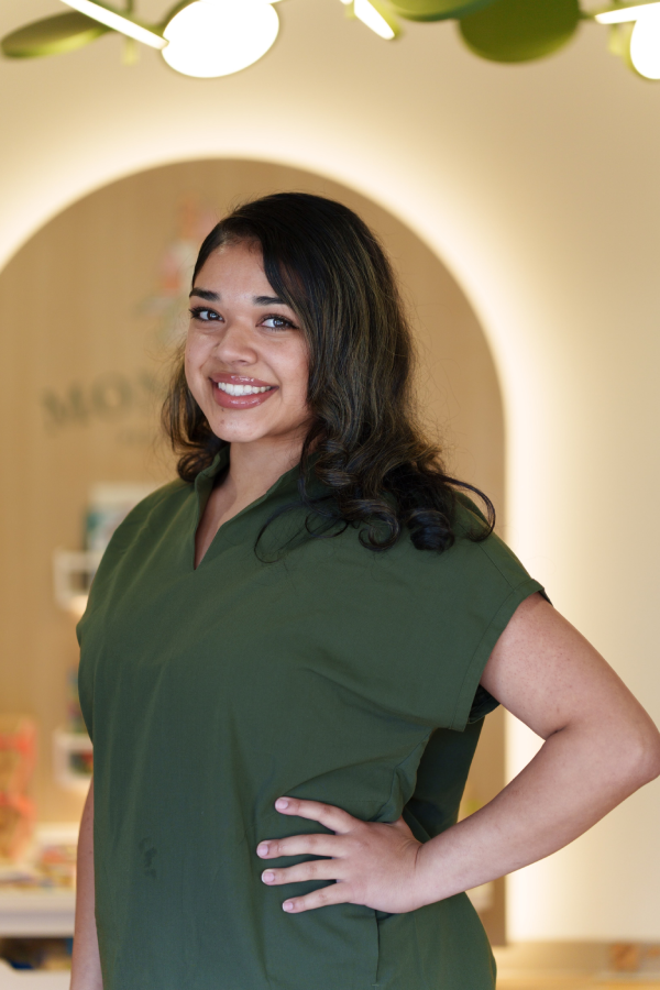 Smiling staff member with long dark curly hair, wearing a green scrub top, standing with hand on hip in front of a wooden archway with a partial "Mo..." sign, at Montclair pediatric dental office.