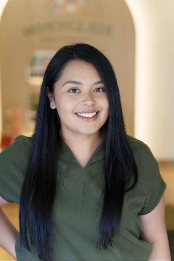 Smiling staff member with dark curled hair, hand on hip, wearing a green scrub top, standing in front of a wooden archway near a green leaf light fixture.